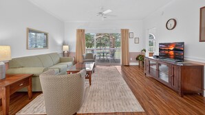 A living room with a green sofa, a patterned armchair, a wooden coffee table, a TV on a wooden cabinet, and a view of a balcony with outdoor furniture.