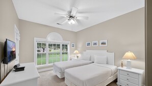 A bedroom with two beds, a ceiling fan, a desk with a TV, and a window with a view of a green lawn.