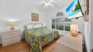 A bedroom with a bed, a nightstand, a dresser, a ceiling fan, a TV mounted on the wall, and a window with a view of greenery. - Morning sun | golf fun | ocean breeze retreat | Magnolia Place 4701-103 (Myrtle Beach)