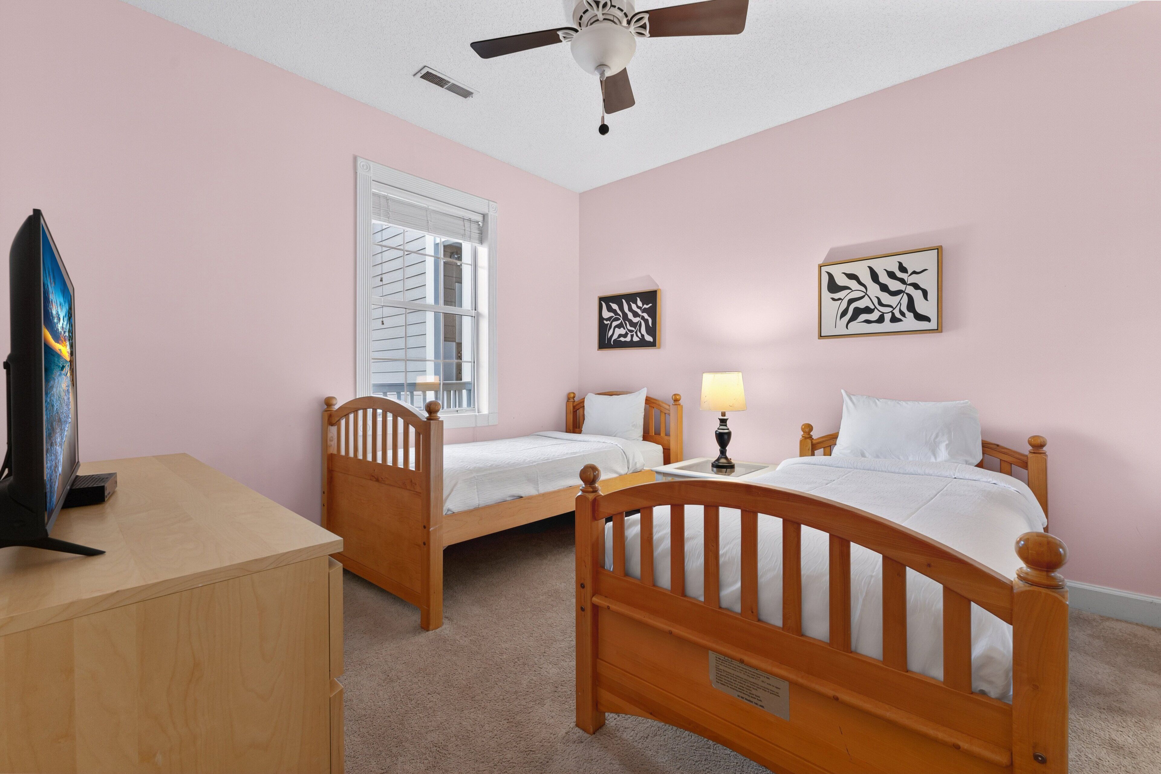 Une chambre à coucher avec deux lits en bois, un ventilateur de plafond, une fenêtre avec des stores et un bureau équipé d’un écran d’ordinateur.