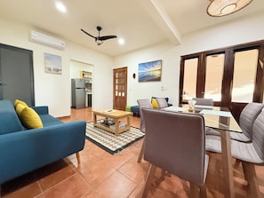 A living room with a blue sofa, a grey armchair, a coffee table, and a dining table with chairs. There is a kitchen area with a refrigerator and a dining table.