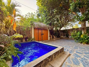 A swimming pool with a stone border, surrounded by tropical plants and a wooden structure with a thatched roof.