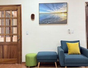 Una habitación con puerta de madera, un cuadro enmarcado de una puesta de sol sobre el océano, un sofá azul con un cojín amarillo y un otoman verde.