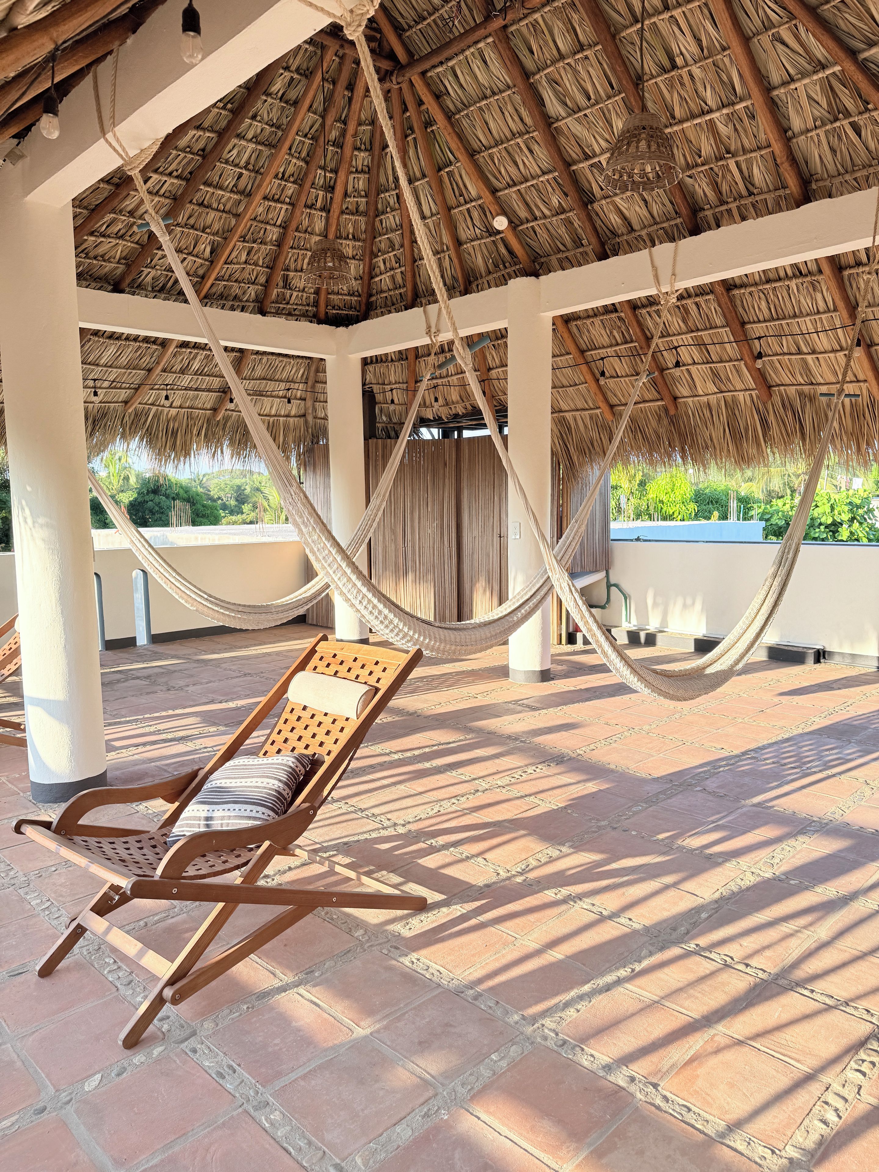 A hammock under a thatched roof structure with a wooden lounge chair and tiled flooring.