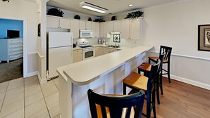 A modern kitchen with white cabinets, a white refrigerator, a microwave, and a stove. There are two wooden bar stools and a white countertop. - Magnolia Pointe Retreat Near the Beach | Magnolia Pointe Magnolia Lake 4812-201 (Myrtle Beach)