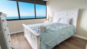 A bedroom with a bed, a dresser, a nightstand, and a window with a view of the ocean.