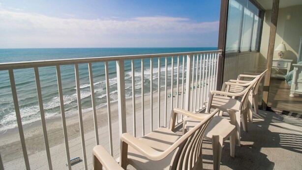 A balcony with a view of the ocean, featuring plastic chairs and a glass railing.