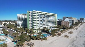 Un grand hôtel à plusieurs étages, doté de balcons, situé près d’une plage, avec une piscine et des palmiers.