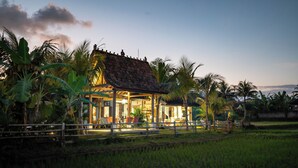 A traditional wooden house with a tiled roof, surrounded by lush greenery and a rice field.