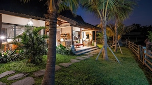 A well-lit house with a stone pathway leading to a porch, surrounded by palm trees and greenery.