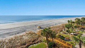 A beach view with a wooden walkway, palm trees, and beach huts.