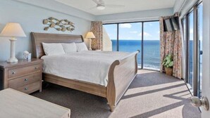 A hotel room with a bed, a nightstand, a lamp, a ceiling fan, a view of the ocean, and a large window. - Savoy 3BR Oceanfront Condo + Gym & Beach Access! | Savoy PH (Myrtle Beach)