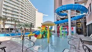 A hotel pool area with a water slide, a colorful play structure, and a large building in the background. - Savoy 3BR Oceanfront Condo + Gym & Beach Access! | Savoy PH (Myrtle Beach)