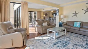 A living room with a beige sofa, a leather armchair, and a coffee table. There is a balcony with curtains, a dining area in the background, and star decorations on the wall. - Savoy 3BR Oceanfront Condo + Gym & Beach Access! | Savoy PH (Myrtle Beach)