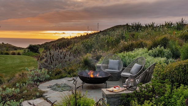Une terrasse avec un foyer, des fauteuils en osier et une vue sur l’océan au coucher du soleil.