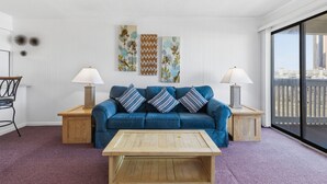 A living room with a blue sofa, two side tables with lamps, a wooden coffee table, and three wall-mounted artworks. - Cozy Condo | Stunning Ocean View in Myrtle Beach | A Place at the Beach IV G231 (Myrtle Beach)