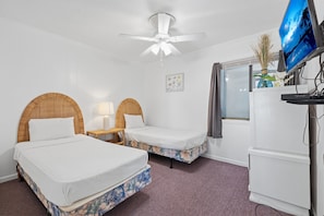Une chambre avec deux lits, un ventilateur de plafond, une télévision, une fenêtre et une petite table avec une lampe.