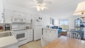 A modern kitchen with white appliances, a dining area with a wooden table, and a living room with a blue sofa. - Cozy Condo | Stunning Ocean View in Myrtle Beach | A Place at the Beach IV G231 (Myrtle Beach)