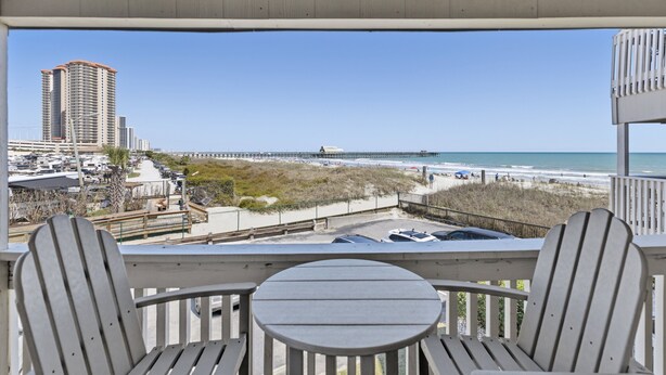 A balcony with two wooden chairs and a small table, overlooking a beach with a boardwalk and high-rise buildings in the distance. - Cozy Condo | Stunning Ocean View in Myrtle Beach | A Place at the Beach IV G231 (Myrtle Beach)