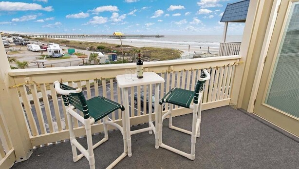 A balcony with two white chairs and a small table, overlooking a beach and a pier.