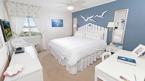 A bedroom with a bed, a desk, a chair, a ceiling fan, a window with curtains, a painting, and a wall-mounted mirror.