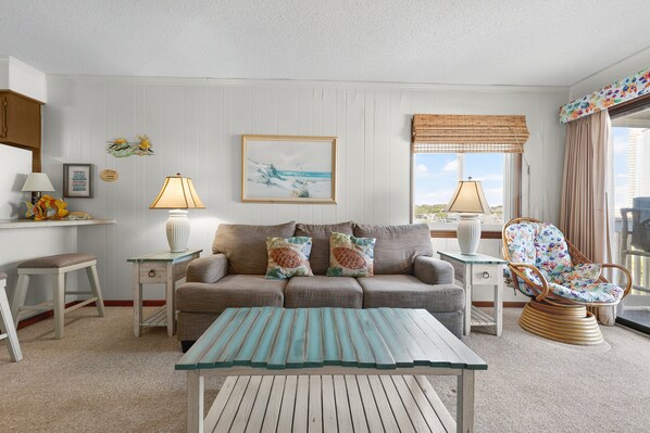 A living room with a sofa, two side tables, a wicker chair, a glass coffee table, and a view of the outdoors.