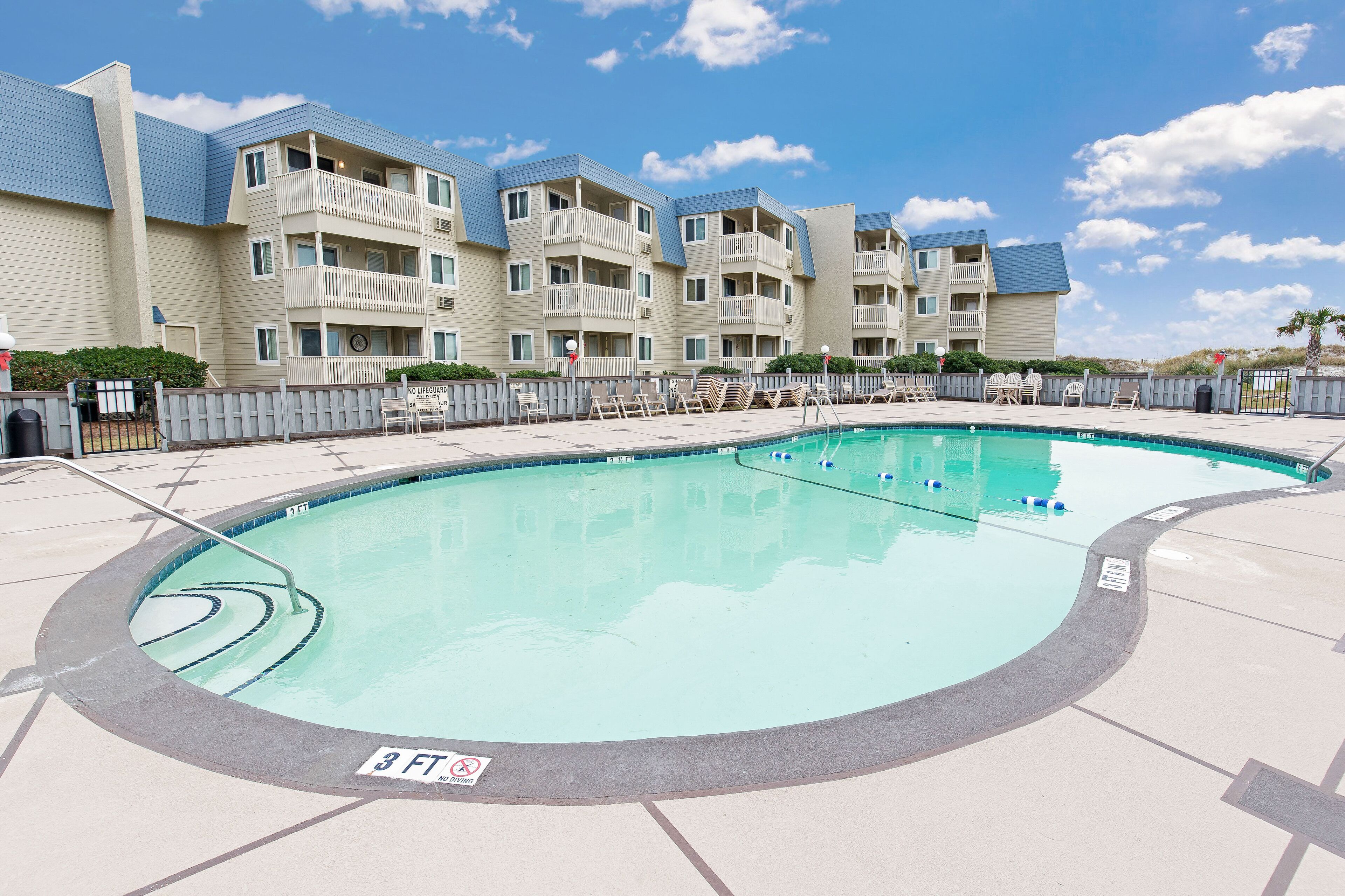 A swimming pool with a 3 FT depth sign, surrounded by a fence, and adjacent to a multi-story building with balconies.