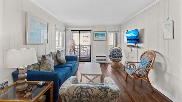 A living room with a blue sofa, a white coffee table, and a wicker chair. There is a television mounted on the wall and a window with a view of the outside. - A Place at the Beach V A204, Ocean View Comfort Awaits You in Myrtle Beach (Myrtle Beach)