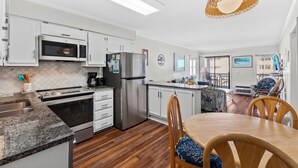 A modern kitchen with white cabinets, stainless steel appliances, a dining area with wooden chairs, and a large window offering a view of the city. - A Place at the Beach V A204, Ocean View Comfort Awaits You in Myrtle Beach (Myrtle Beach)