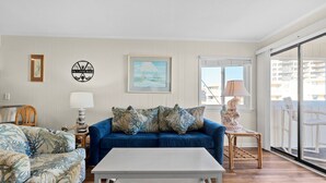A living room with a blue sofa, a matching armchair, a white coffee table, and a view of the balcony through a large window. - A Place at the Beach V A204, Ocean View Comfort Awaits You in Myrtle Beach (Myrtle Beach)