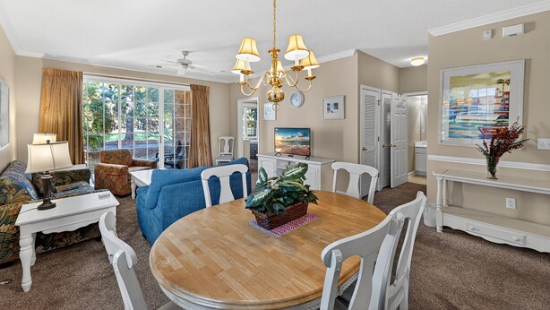 A dining area with a round wooden table, white chairs, and a chandelier. There is a blue sofa and a television in the background.