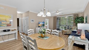A dining area with a round wooden table, white chairs, a chandelier, and a view of the outdoors through large windows. - Arbor Trace 322, Condo by the Greens, Relaxing Golf Views Await (North Myrtle Beach)