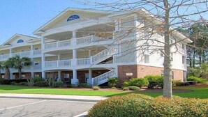 A multi-story hotel with balconies, a parking lot, and well-maintained landscaping. - Arbor Trace 322, Condo by the Greens, Relaxing Golf Views Await (North Myrtle Beach)