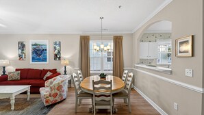 A dining area with a round wooden table, chairs, and a chandelier. There is a red sofa and a white coffee table in the living area. - Arbor Trace 724, Tranquil Golf Views, Walk to Tee Time Daily (North Myrtle Beach)