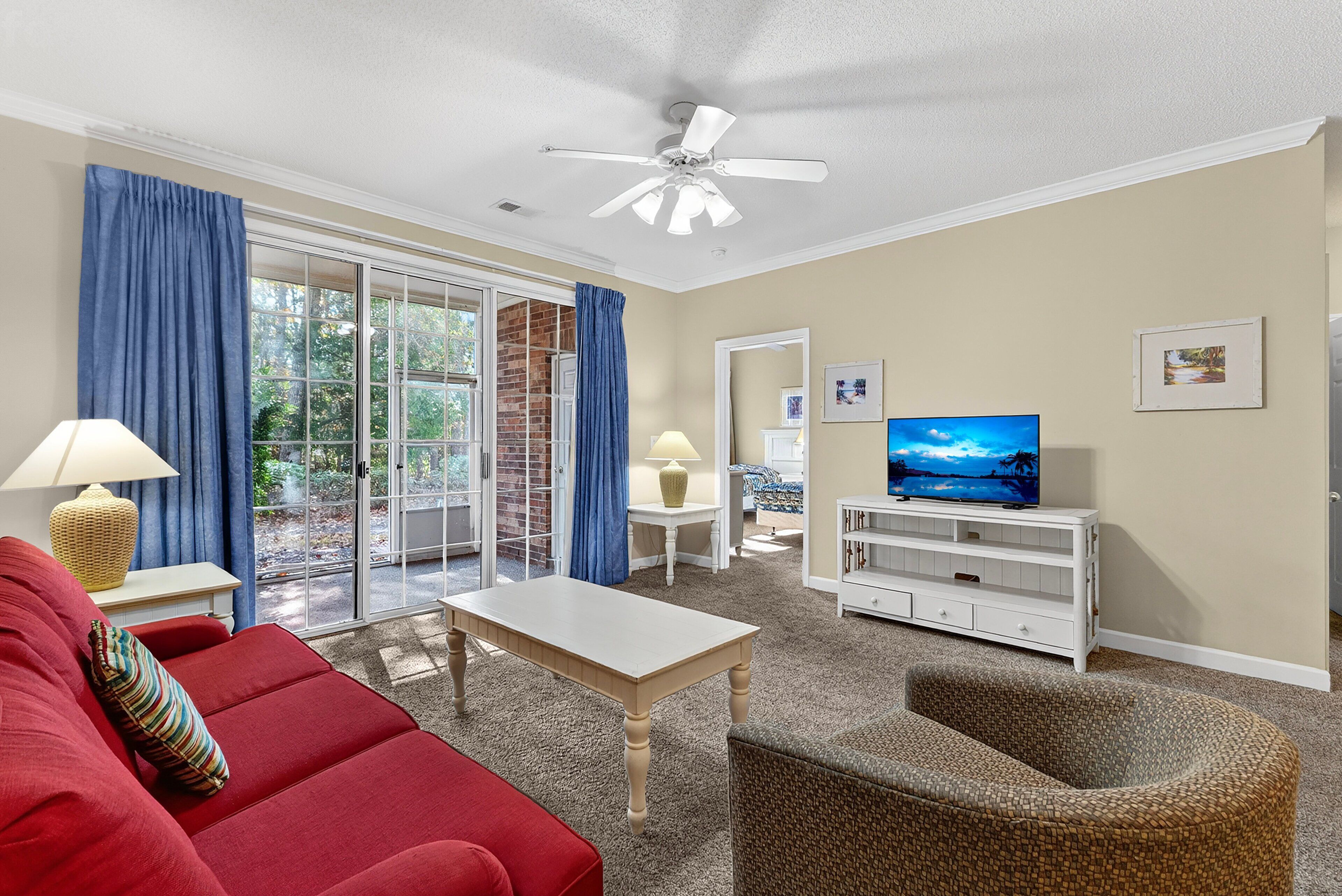 A living room with a red sofa, a white coffee table, a wicker chair, a television on a white stand, and a ceiling fan.