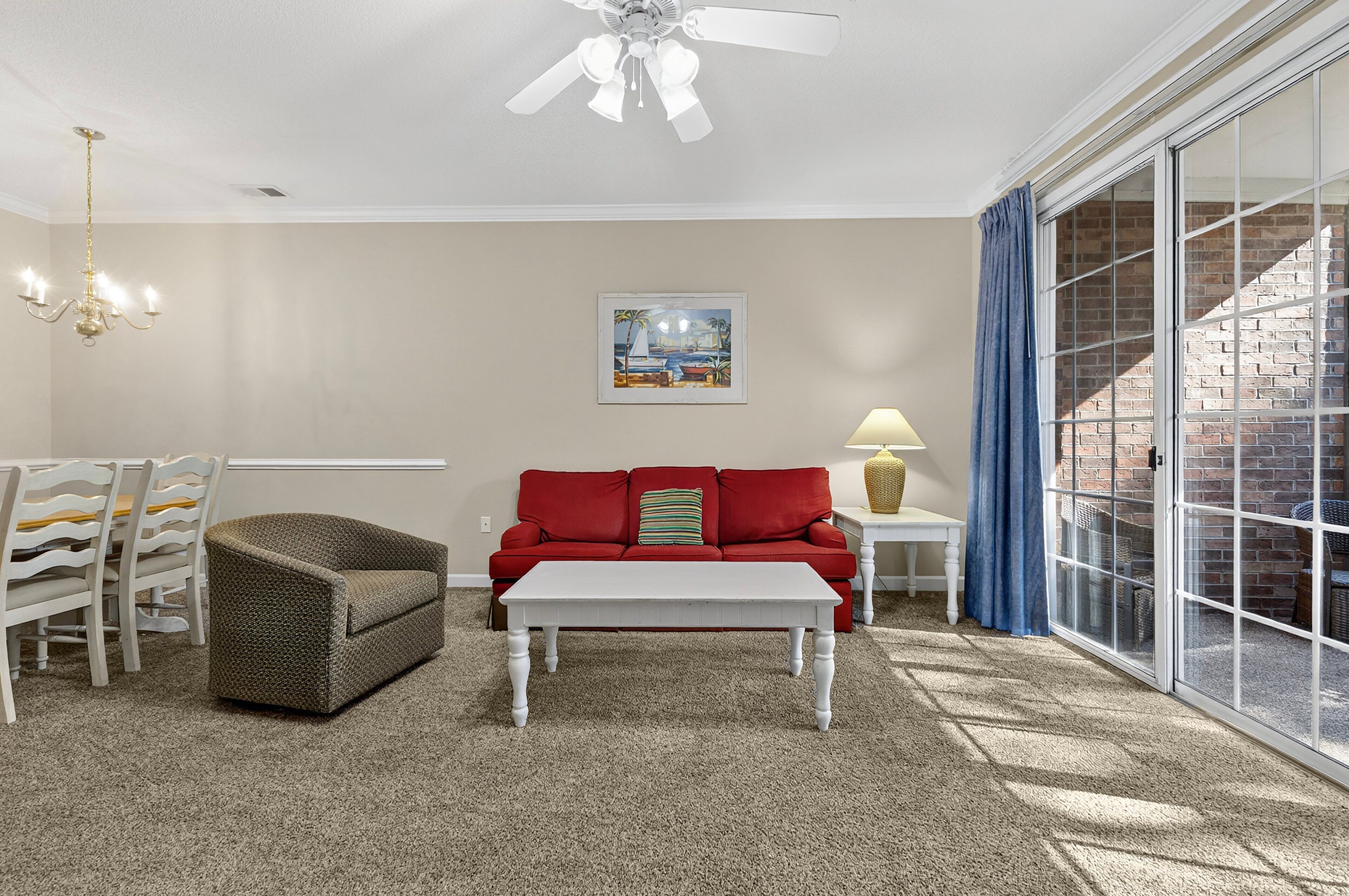 A living room with a red sofa, white coffee table, and a chandelier. There is a painting on the wall and a window with blue curtains.