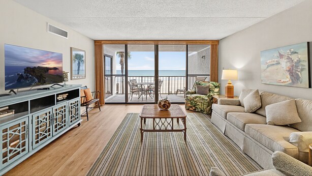 A modern living room with a TV, a sofa, a coffee table, and a view of the ocean through large glass doors.