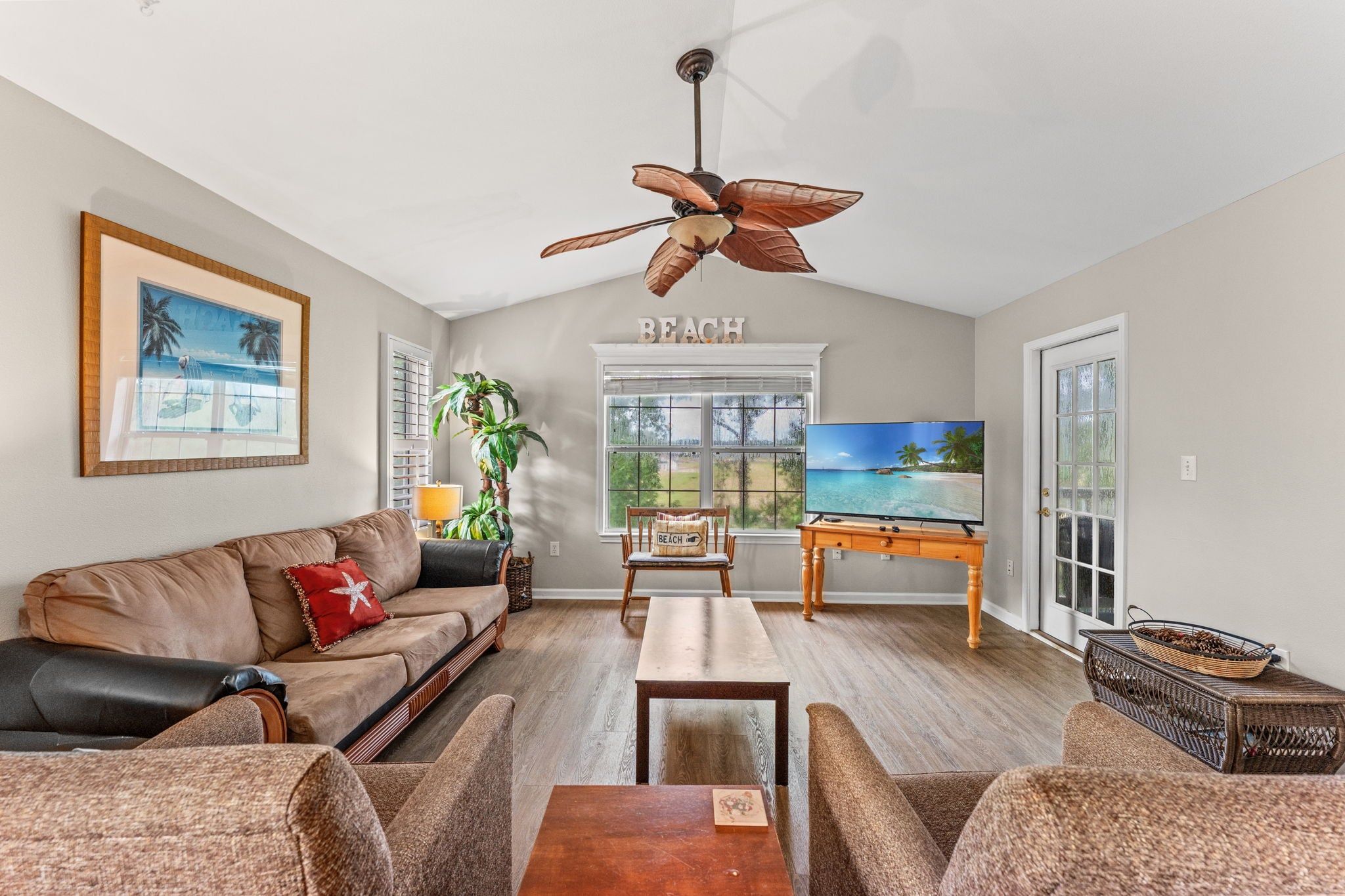 A living room with a sofa, a coffee table, a TV, a ceiling fan, a window with a view, and a wall with a 'BEACH' sign.
