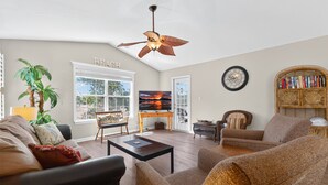 A living room with a ceiling fan, a television, a bookshelf, and a large window. - River Oaks Getaway | Golf, Beach, Fun – All in One! | Fairways 26E (Myrtle Beach)