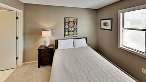 A bedroom with a bed, a nightstand with a lamp, a framed floral artwork, and a window with blinds. - Beach Cottage Retreat | Relax by the Shore & Golf | GC 6 (Myrtle Beach)