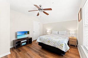 Quarto com cama, ventoinha no teto, televisão, mesa de cabeceira e janela com persianas.