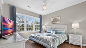 A bedroom with a bed, nightstands, a TV, a ceiling fan, and a window with curtains.