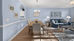 A dining area with a wooden table, chairs, and a chandelier. There is a blue sofa with a patterned rug in the living area.