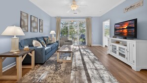 A living room with a blue sofa, a coffee table, a TV on a white cabinet, and a large window with curtains.