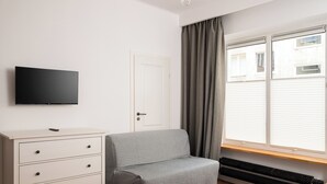 Makuuhuoneessa on sänky, lipasto, harmaa sohva, seinälle asennettu televisio ja ikkuna, jossa on verhot.