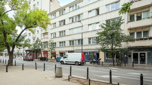 Useita kerroksia sisältävä rakennus, jonka julkisivu on valkoinen. Valkoinen pakettiauto on pysäköitynä, ja jalkakäytävällä on moottoripyörä.