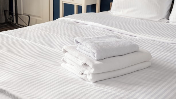 Een net opgemaakt bed met witte lakens en een stapel opgevouwen handdoeken.