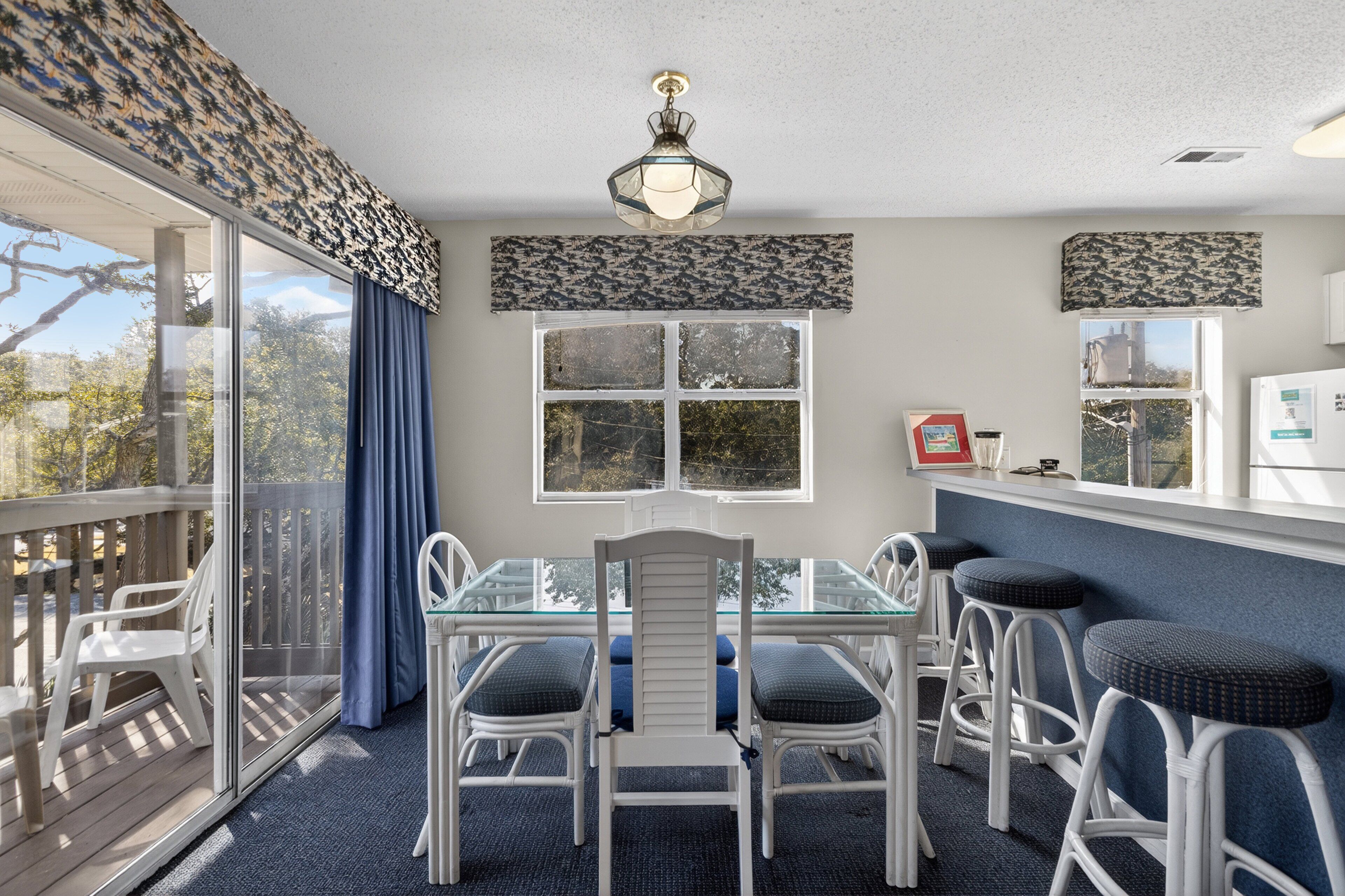 Un coin repas avec une table en verre et des chaises blanches, un rideau bleu et une vue sur les arbres par la fenêtre.