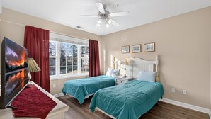 Ein Schlafzimmer mit zwei Betten, einem Fernseher, einem Deckenventilator und einem Fenster mit Vorhängen.