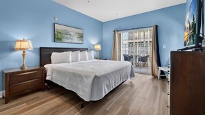 Une chambre à coucher comprenant un lit, des tables de chevet, un ventilateur de plafond, un tableau, une fenêtre avec des rideaux et une vue sur un balcon.